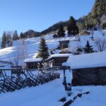 Schneebedeckte Holzhütten und Zäune an einem Hang, umgeben von Bäumen und Bergen unter einem klaren blauen Himmel.