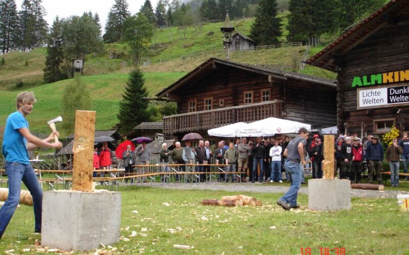 Zwei Personen nehmen an einem Holzhackwettbewerb im Freien teil, während eine Menschenmenge in der Nähe von Holzgebäuden und Bäumen im Hintergrund zusieht.