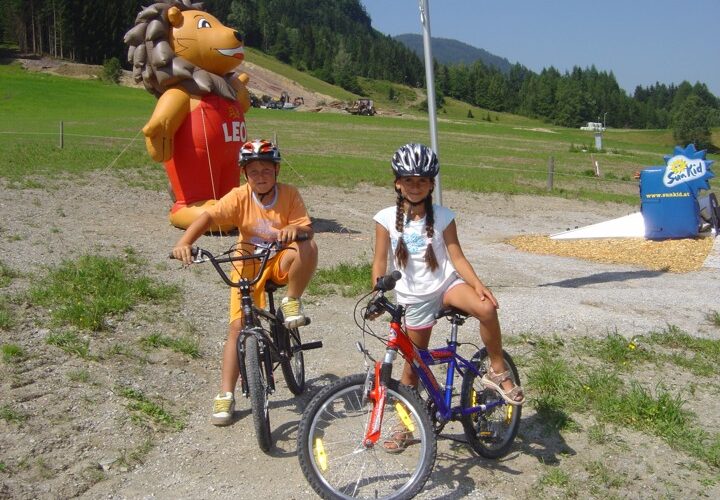 Zwei Kinder mit Helmen sitzen auf Fahrrädern auf einem Schotterweg mit einem großen aufblasbaren Löwen und grasbewachsenen Hügeln im Hintergrund.