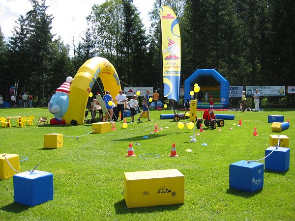 Kinderspielplatz im Freien mit aufblasbaren Strukturen, bunten Blöcken, Kegeln und Luftballons auf einer Wiese, wo Erwachsene und Kinder an Aktivitäten teilnehmen.