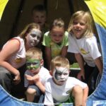 Sechs Kinder mit Gesichtsbemalung posieren an einem sonnigen Tag gemeinsam am Eingang eines gelb-blauen Zeltes.