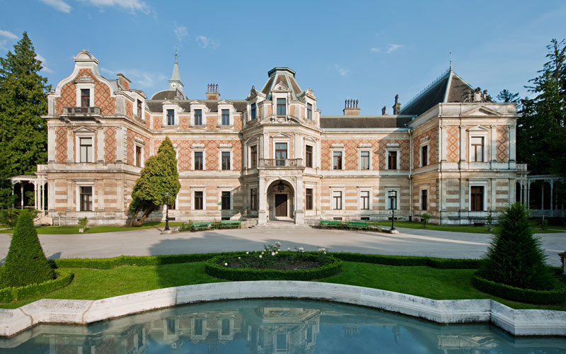 Ein großes, verziertes historisches Herrenhaus mit gemustertem Mauerwerk steht hinter einem gepflegten Garten und einem geschwungenen, spiegelnden Pool an einem klaren Tag.