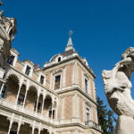 Verziertes historisches Gebäude mit spitzem Turm und verschlungenen Balkonen, neben einer weißen Steinstatue, vor einem klaren blauen Himmel.