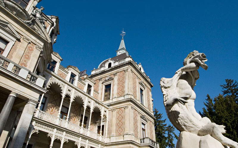 Verziertes historisches Gebäude mit spitzem Turm und verschlungenen Balkonen, neben einer weißen Steinstatue, vor einem klaren blauen Himmel.