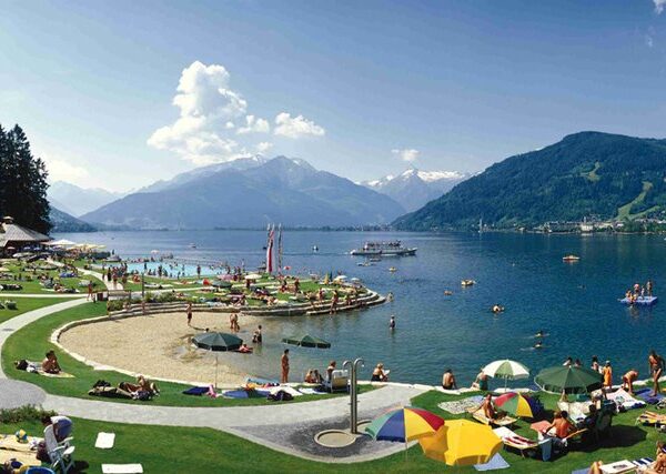 Die Menschen entspannen sich auf Gras- und Sandflächen an einem See mit Bergen im Hintergrund; einige schwimmen oder fahren mit Tretbooten, während andere sich unter Sonnenschirmen sonnen.