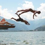 Zwei Personen sitzen auf einem Steg, während zwei andere in einen See springen. Im Hintergrund sind Berge und ein bewölkter Himmel zu sehen.