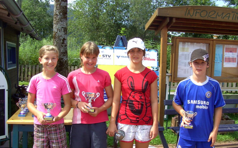 Vier Kinder stehen im Freien und halten Trophäen, im Hintergrund sind Informationstafeln und Grünpflanzen zu sehen.