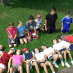 Eine Gruppe von Kindern posiert auf einer Wiese; einige stehen oder knien mit Trophäen, andere liegen lächelnd nebeneinander, in legerer Sportkleidung.