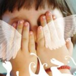 Ein Kind bedeckt sein Gesicht mit beiden Händen; weiße illustrierte Flügel und dekorative Wirbel sind über das Bild gelegt.