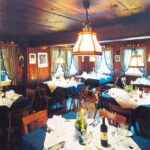 Ein gemütlicher, holzgetäfelter Speisesaal mit mehreren gedeckten Tischen, Holzstühlen, großen Fenstern mit Vorhängen und Hängelampen.