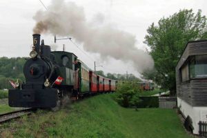 Eine schwarze Dampflokomotive zieht eine Reihe von bunten Personenwagen über eine grasbewachsene Bahnstrecke, die weißen Rauch ausstößt, mit Bäumen und einem Gebäude in der Nähe.
