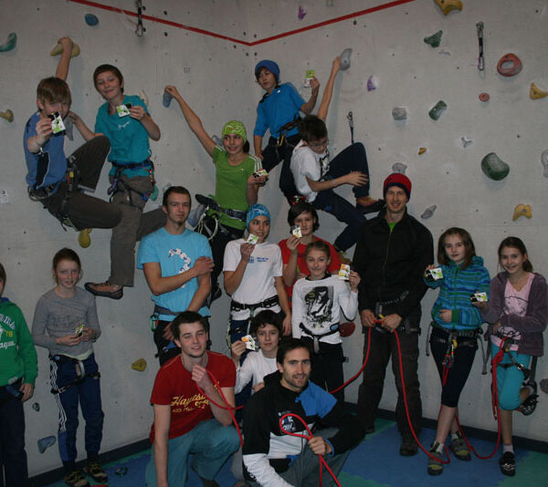 Eine Gruppe von Kindern und Erwachsenen posiert vor einer Indoor-Kletterwand, einige tragen Klettergurte und Helme, halten Karten und Kletterausrüstung.