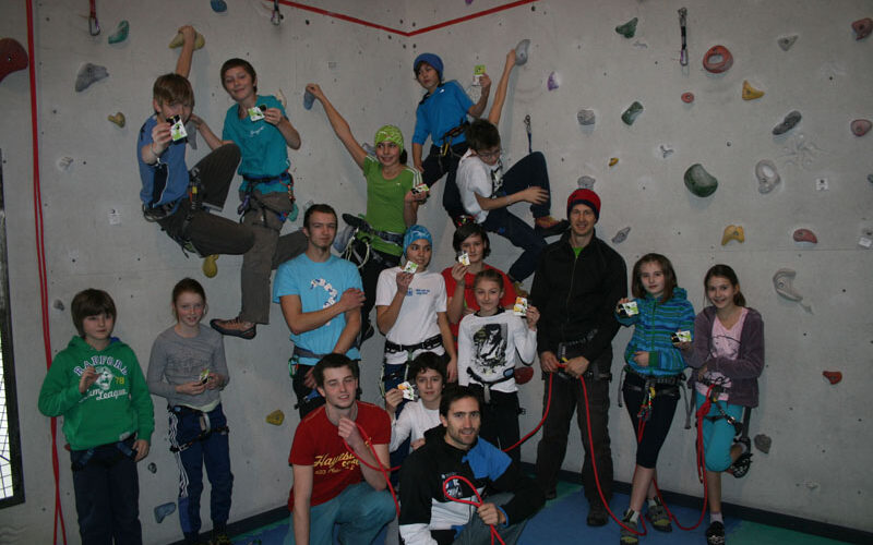 Eine Gruppe von Kindern und Erwachsenen posiert vor einer Indoor-Kletterwand, einige tragen Klettergurte und Helme, halten Karten und Kletterausrüstung.