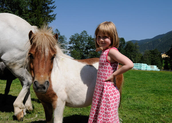 Ein junges Mädchen in einem rosa Kleid steht neben einem kleinen braun-weißen Pony auf einer Wiese mit Bäumen und Bergen im Hintergrund.
