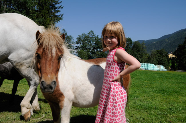 Ein junges Mädchen in einem rosa Kleid steht neben einem kleinen braun-weißen Pony auf einer Wiese mit Bäumen und Bergen im Hintergrund.