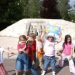 Eine Gruppe von sieben Kindern posiert und lächelt an einem sonnigen Tag vor einer Sand- oder Steinskulptur im Freien.