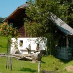 Ein rustikales weißes Holzhaus mit einem Wasserrad, umgeben von grünem Gras, Bäumen und einem Holzzaun an einem sonnigen Tag.