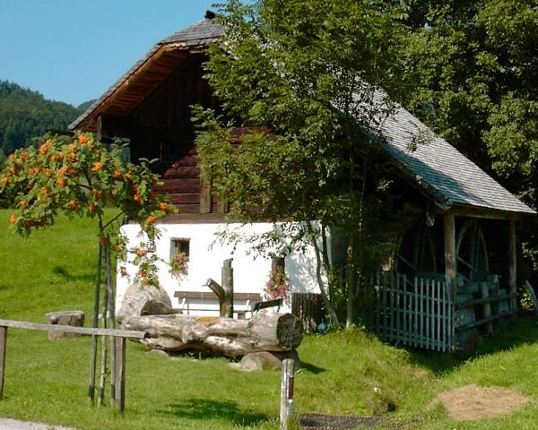 Ein rustikales weißes Holzhaus mit einem Wasserrad, umgeben von grünem Gras, Bäumen und einem Holzzaun an einem sonnigen Tag.