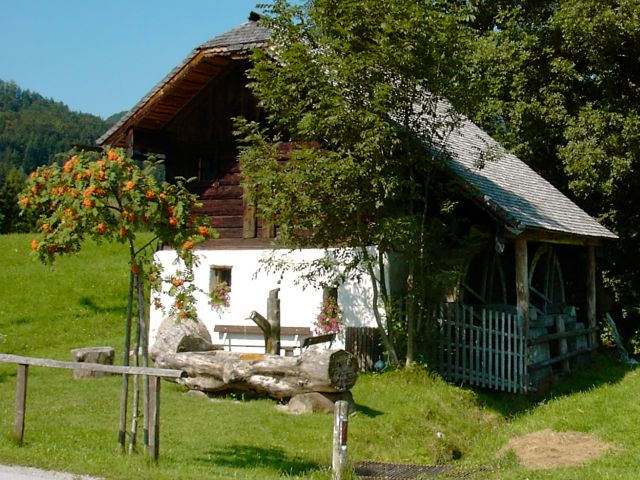 Ein rustikales weißes Holzhaus mit einem Wasserrad, umgeben von grünem Gras, Bäumen und einem Holzzaun an einem sonnigen Tag.