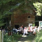 Eine Gruppe von Menschen sitzt an Picknicktischen vor einer rustikalen Holzhütte in einem Waldgebiet, während das Sonnenlicht durch die Bäume fällt.