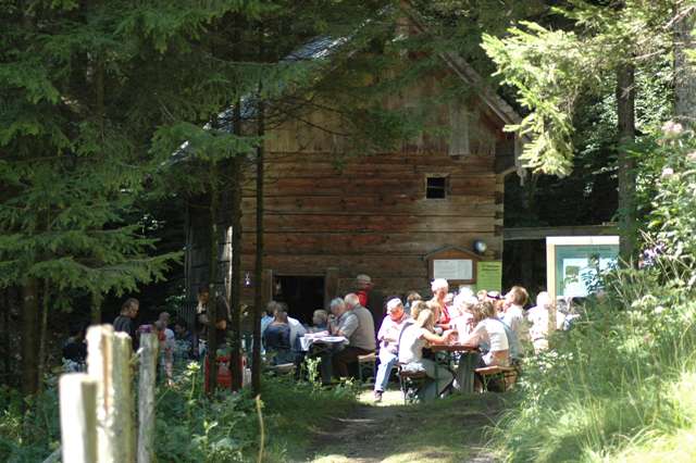 Eine Gruppe von Menschen sitzt an Picknicktischen vor einer rustikalen Holzhütte in einem Waldgebiet, während das Sonnenlicht durch die Bäume fällt.