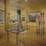 Eine Museumsgalerie mit Vitrinen, in denen Artefakte, Werkzeuge und Skulpturen ausgestellt sind; an den gelben Wänden hängen Gemälde; Holzfußboden und Deckenleuchten sind zu sehen.