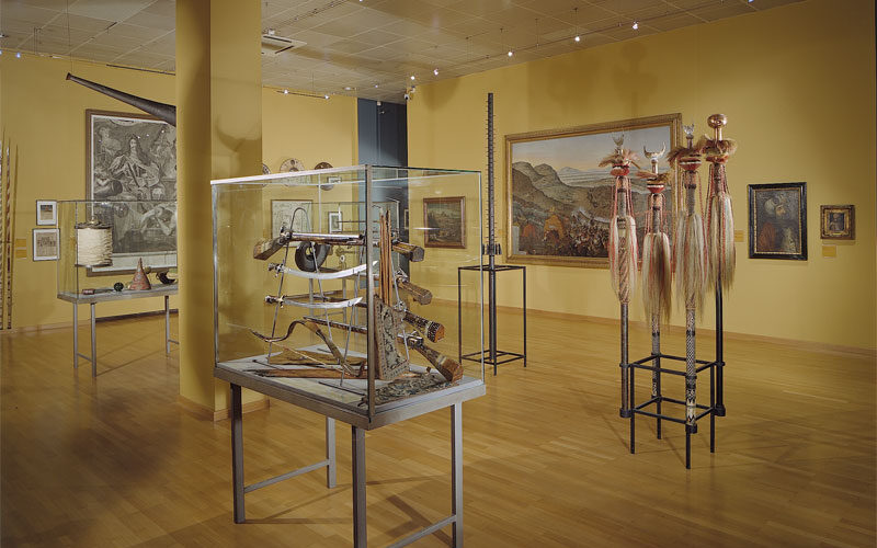 Eine Museumsgalerie mit Vitrinen, in denen Artefakte, Werkzeuge und Skulpturen ausgestellt sind; an den gelben Wänden hängen Gemälde; Holzfußboden und Deckenleuchten sind zu sehen.