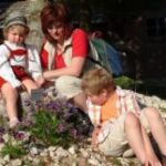 Ein Erwachsener und zwei Kinder sitzen auf Steinen im Freien in der Nähe von lila Blumen, mit einem Holzgebäude und Grünpflanzen im Hintergrund.