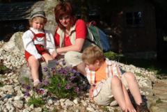 Ein Erwachsener und zwei Kinder sitzen auf Steinen im Freien in der Nähe von lila Blumen, mit einem Holzgebäude und Grünpflanzen im Hintergrund.