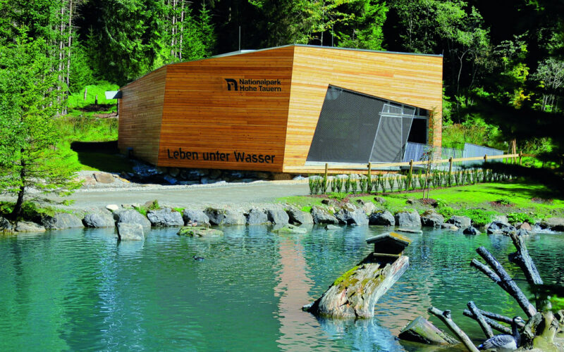 Ein modernes Holzgebäude mit der Aufschrift "Leben unter Wasser" steht an einem klaren, grünlichen Teich, umgeben von Felsen, Baumstämmen und dichtem Wald.
