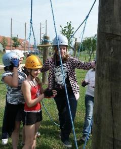 Vier Personen mit Helmen und Sicherheitsausrüstung stehen im Freien in einem Hochseilgarten und interagieren mit Seilen und Holzpfosten.