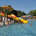 Menschen genießen einen sonnigen Tag in einem Freibad mit einer gelben und orangefarbenen Wasserrutsche. Einige schwimmen, während andere darauf warten, dass sie mit der Rutsche an der Reihe sind. Bäume und Sitzgelegenheiten am Beckenrand sind zu sehen.