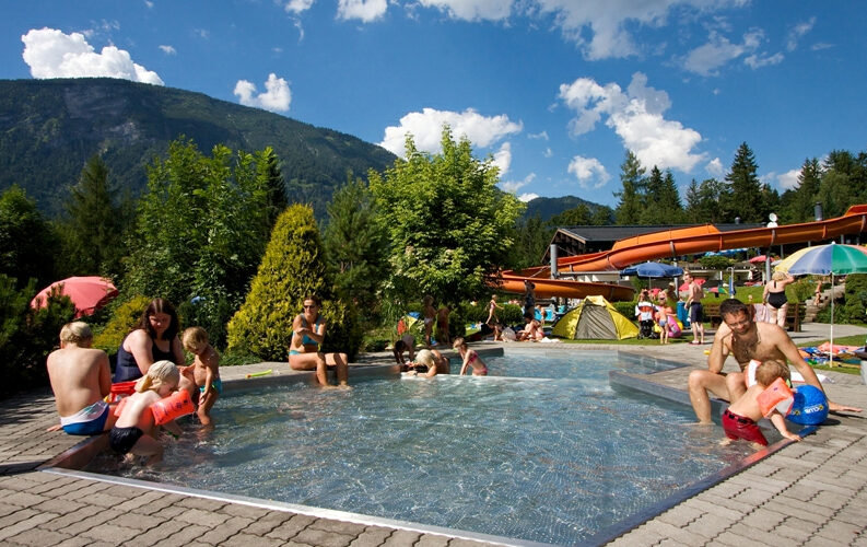 Außenpool mit Kindern und Erwachsenen im flachen Wasser, umgeben von Grün, Rutschen und Bergen unter einem teilweise bewölkten Himmel.