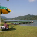 Menschen entspannen sich auf Liegestühlen unter einem Sonnenschirm in der Nähe eines Stegs an einem See, mit Schwimmern im Wasser und grünen Hügeln im Hintergrund.