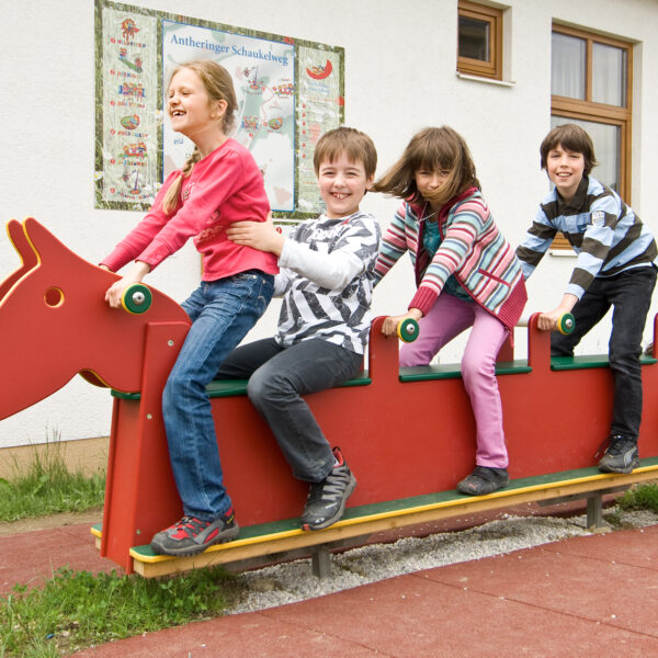 Vier Kinder sitzen lächelnd auf einer roten Spielplatzwippe in Form eines Pferdes, die im Freien vor einem Gebäude steht.