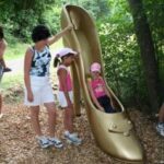 Eine Frau und zwei Kinder stehen im Freien neben einer riesigen goldenen Schuhskulptur mit hohen Absätzen; ein Kind sitzt im Inneren des Schuhs.