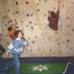 Drei Kinder klettern und spielen an einer Indoor-Kletterwand, unter ihnen liegt eine grüne Matte auf dem gepolsterten Boden.