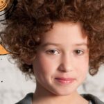 Ein Kind mit lockigem Haar steht vor einer weißen Backsteinmauer, um seinen Kopf herum sind Abbildungen von Uhren, Blumen und Farbspritzern digital hinzugefügt.