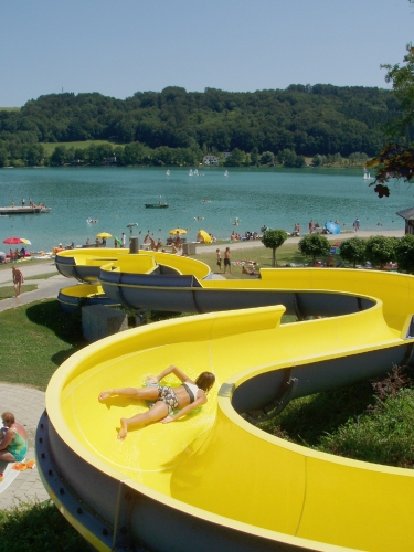 Eine Person rutscht eine leuchtend gelbe Wasserrutsche in der Nähe eines Sees hinunter, während im Hintergrund an einem sonnigen Tag Menschen sonnenbaden, schwimmen und sich entspannen.