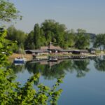 Ein Gebäude am Seeufer mit Anlegestellen und Booten spiegelt sich im ruhigen Wasser, umgeben von grünen Bäumen und einem klaren blauen Himmel.