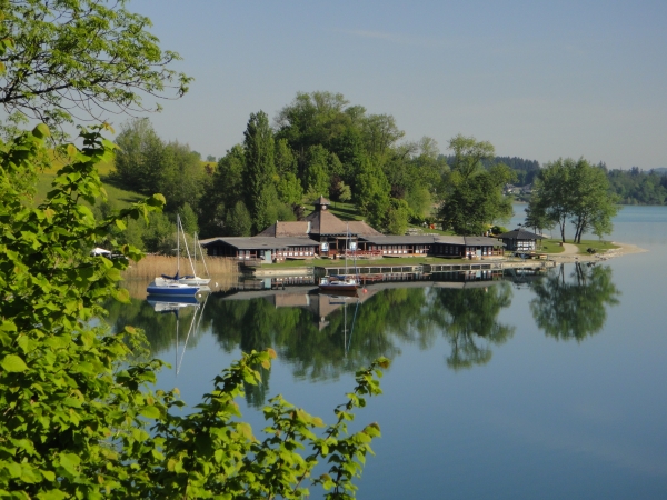 Ein Gebäude am Seeufer mit Anlegestellen und Booten spiegelt sich im ruhigen Wasser, umgeben von grünen Bäumen und einem klaren blauen Himmel.
