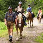 Ein Mann in traditioneller Kleidung führt drei Personen auf einem Pferd über einen Waldweg. Die Reiter tragen Helme und sind leger gekleidet.