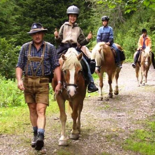 Ein Mann in traditioneller Kleidung führt drei Personen auf einem Pferd über einen Waldweg. Die Reiter tragen Helme und sind leger gekleidet.
