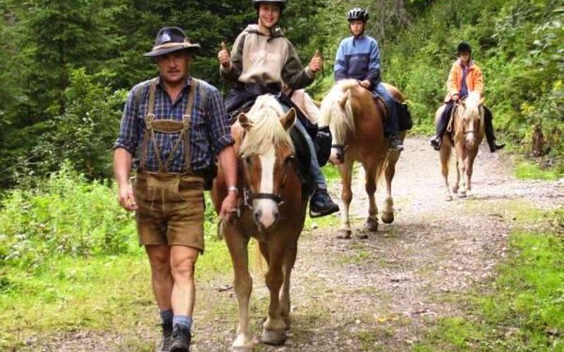 Ein Mann in traditioneller Kleidung führt drei Personen auf einem Pferd über einen Waldweg. Die Reiter tragen Helme und sind leger gekleidet.