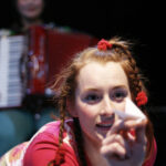 Eine junge Frau mit geflochtenen Haaren und roten Krawatten streckt einen Papiergegenstand in Richtung Kamera, während eine andere Person mit einem Akkordeon im Hintergrund unscharf zu erkennen ist.