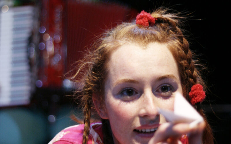 Eine junge Frau mit geflochtenen Haaren und roten Krawatten streckt einen Papiergegenstand in Richtung Kamera, während eine andere Person mit einem Akkordeon im Hintergrund unscharf zu erkennen ist.