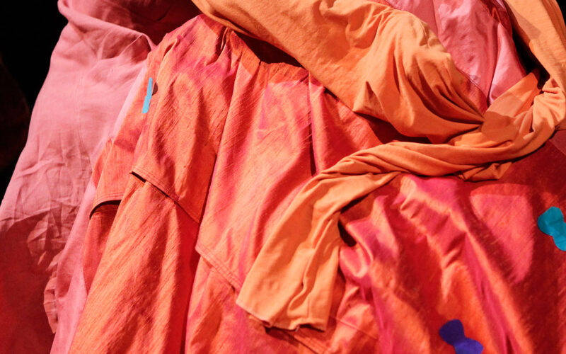 Eine Person in orange-roter Kleidung lehnt ihren Kopf an eine andere Person, die in ähnlich leuchtende Stoffe gehüllt ist, beide auf der Bühne vor einem dunklen Hintergrund.