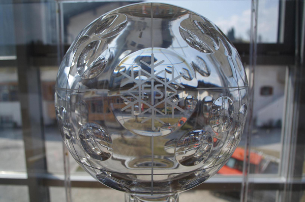 Nahaufnahme einer Trophäe aus Klarglas mit geätztem Schneeflockenmuster, die in einem Innenraum mit Fenstern und einer Außenlandschaft im Hintergrund ausgestellt ist.