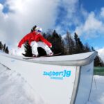 Ein Snowboarder in roter und weißer Ausrüstung gleitet auf einer Schiene in einem verschneiten Skigebiet mit Bäumen und Bergen im Hintergrund.