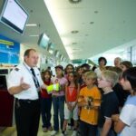 Ein uniformierter Flughafenmitarbeiter erklärt einer Gruppe von Kindern und Erwachsenen, die sich in der Nähe eines Abfertigungsschalters versammelt haben, die Sicherheitsmaßnahmen.